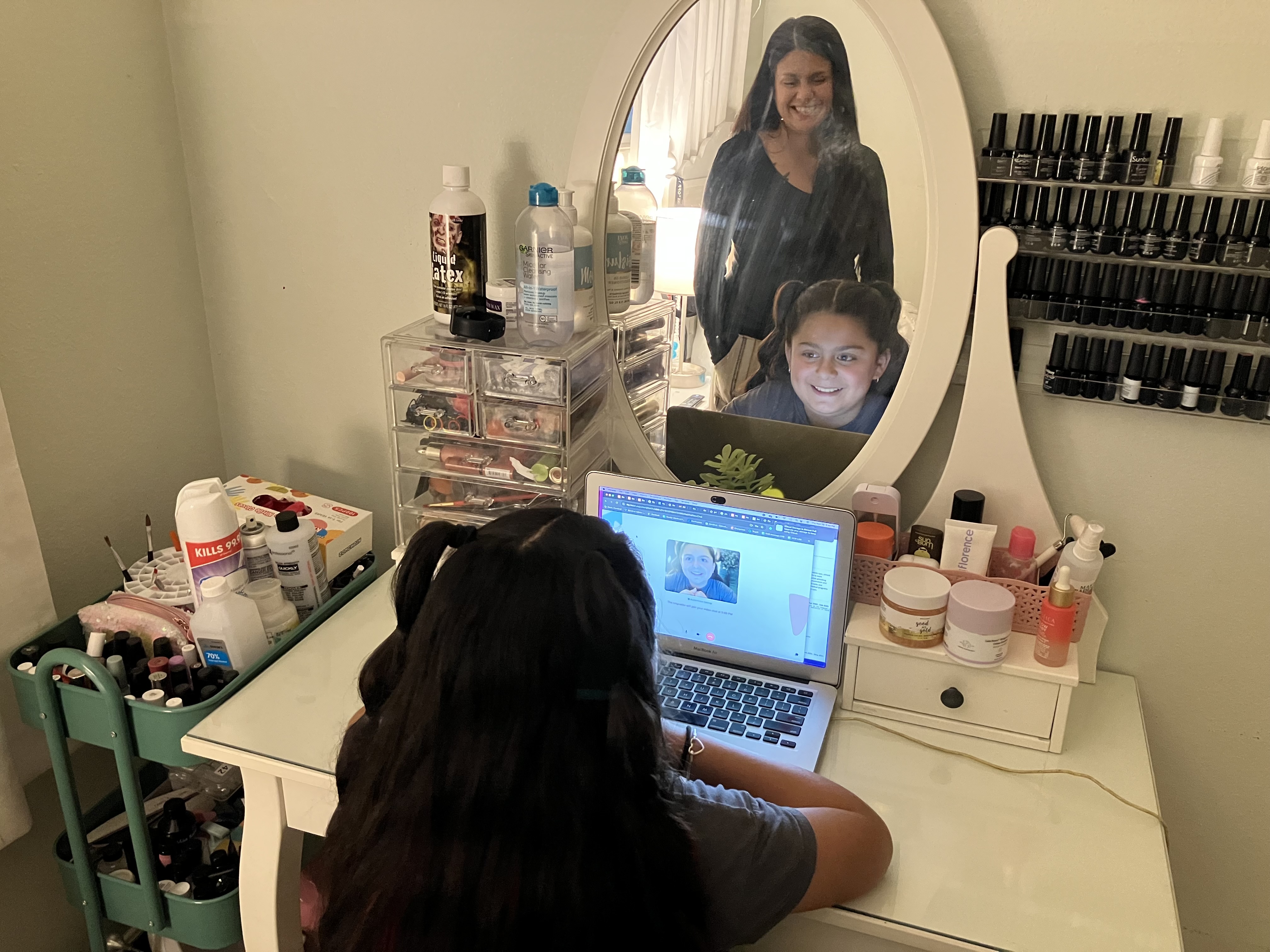 Anjelah Salazar, a young girl, sits at her computer at a desk. A mirror on the desk reflects her mother standing behind her, smiling.
