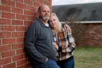 Ron Winters and his wife, Teresa, stand outside their home. Ron leans up against a brick wall, while his wife leans against him and places her hand on his stomach. They both look towards the camera.
