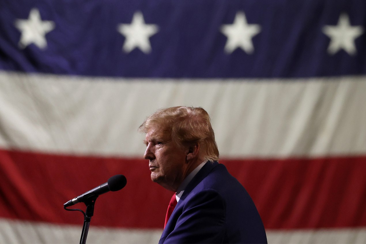 A photo of Former President Donald Trump standing at a microphone with an American flag in the background behind him.