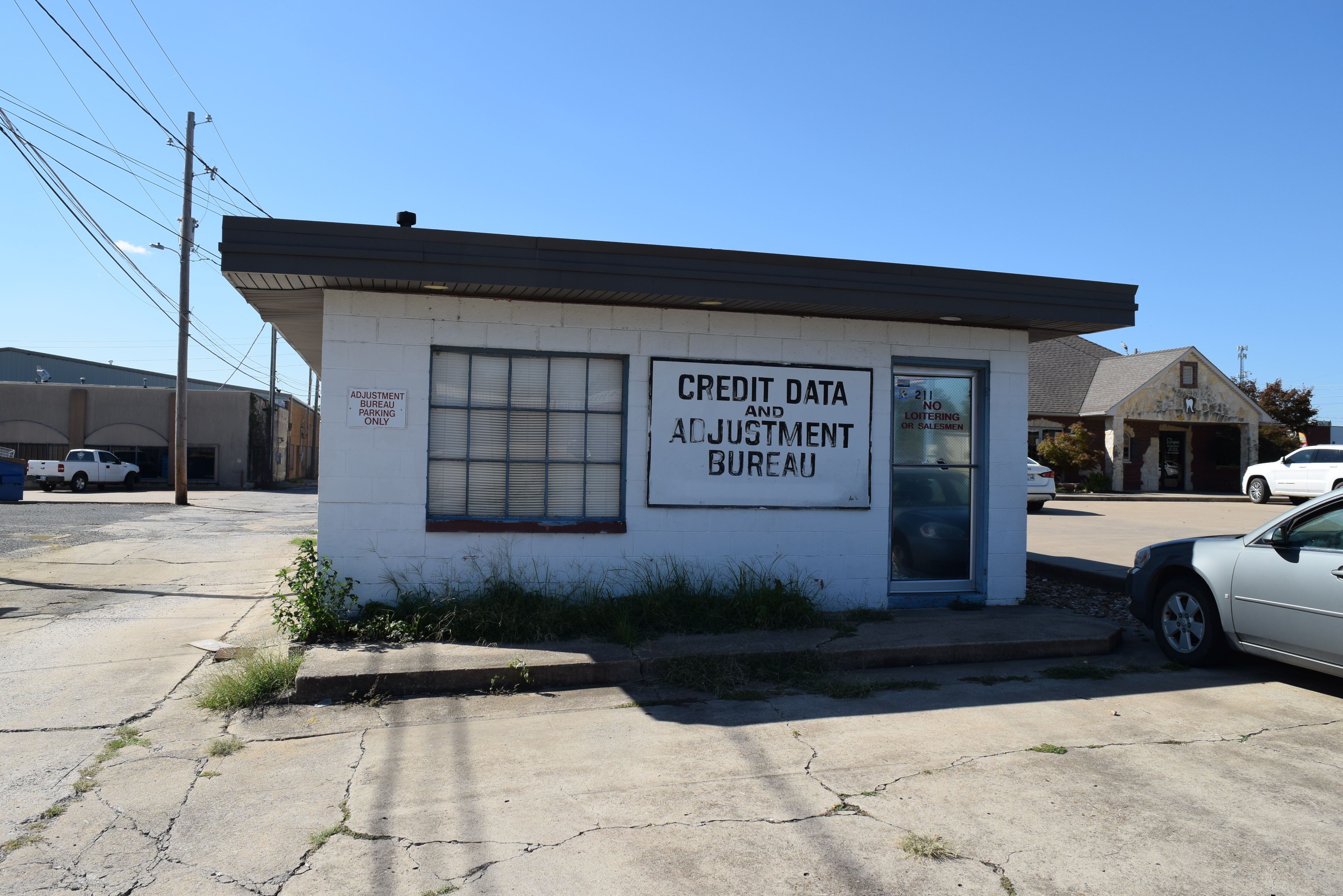 A smalll white building with black roof in a concrete car lot. A sign on the front reads "Credit Data and Adjustment Bureau." 