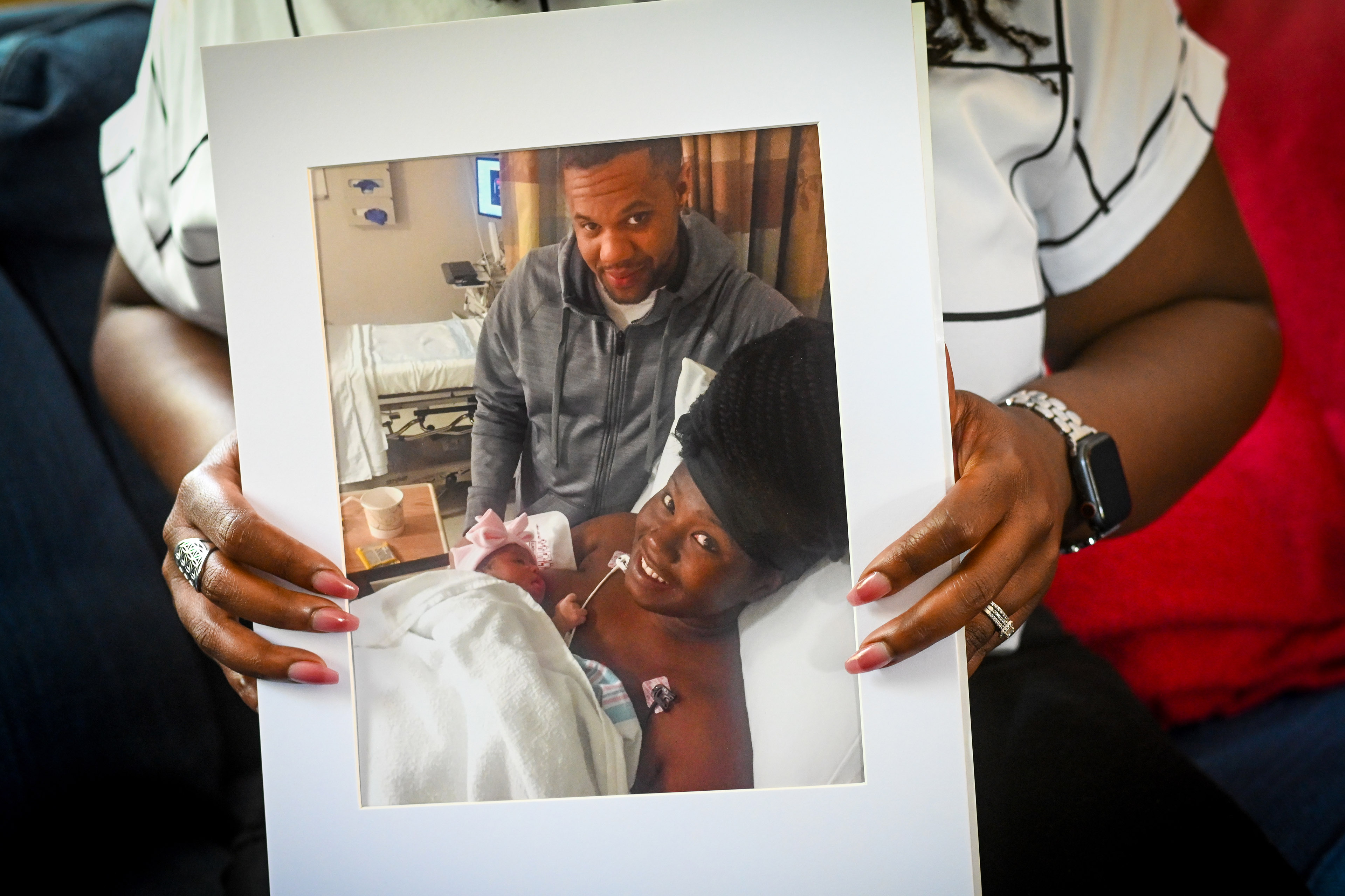 Charity Watkins holds a photo of herself. In the photo, she is lying in a hospital bed with her newborn daughter. Her husband stands beside her.