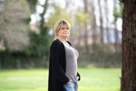A woman with short blonde hair stands in a park outdoors. She is wearing a striped t-shirt, jeans, a black cardigan and a silver cross around her neck