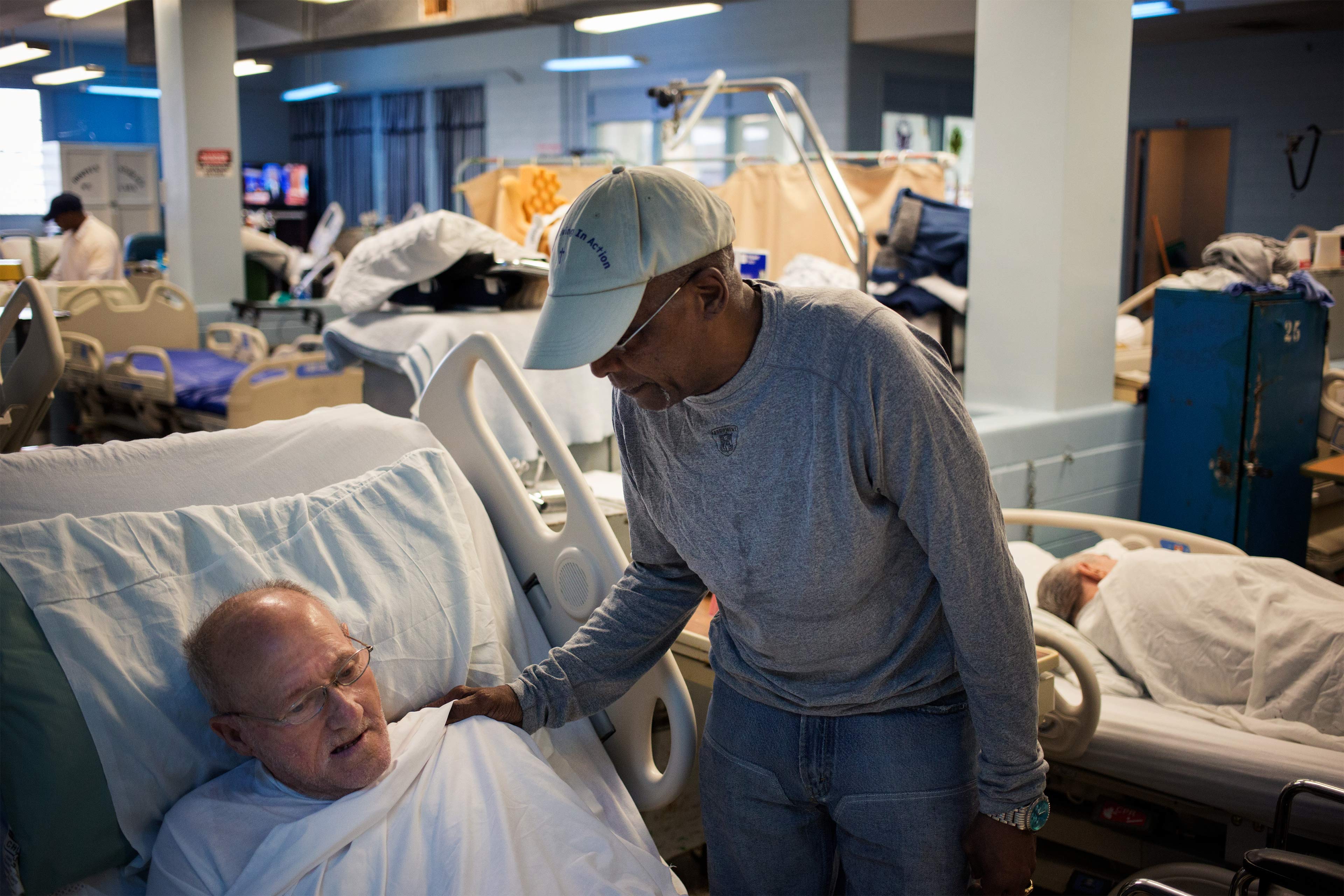 A photo of a younger man wearing touching an older man in a hospital bed on the shoulder.