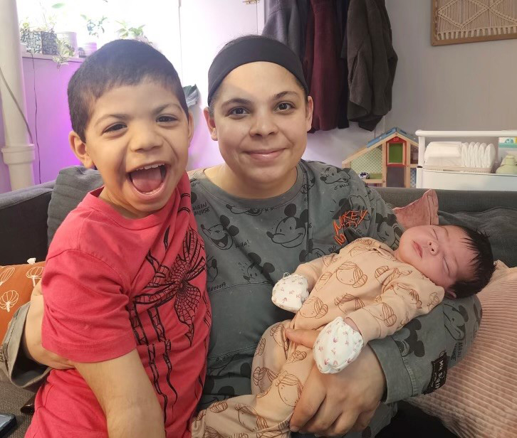 Mary Delgado sits on a couch in her home with one arm around her young son, Joaquin (left). She holds her one-year-hold daughter, Emiliana, in her other arm. Mother and son smile at the camera, while Emiliana sleeps.