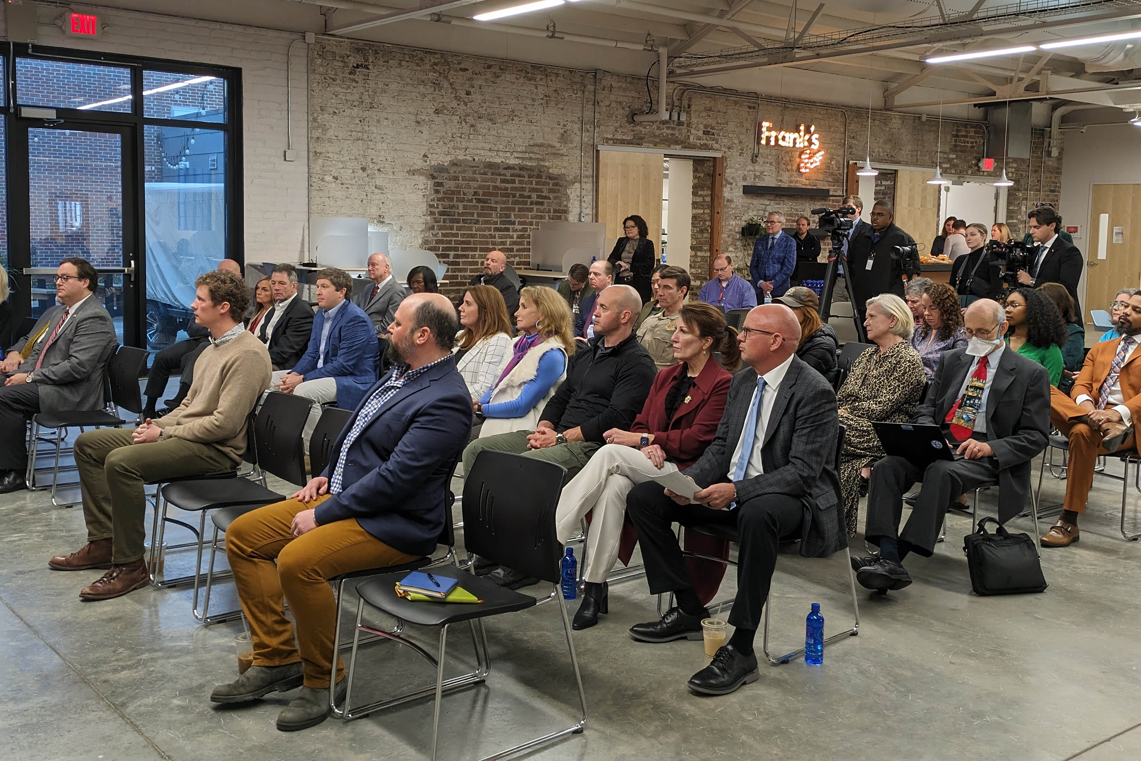 A crowd of about 30 people sit on plastic chairs in rows. They are in a building with a cement floor, exposed brick walls, and crisp, modern lighting overhead.