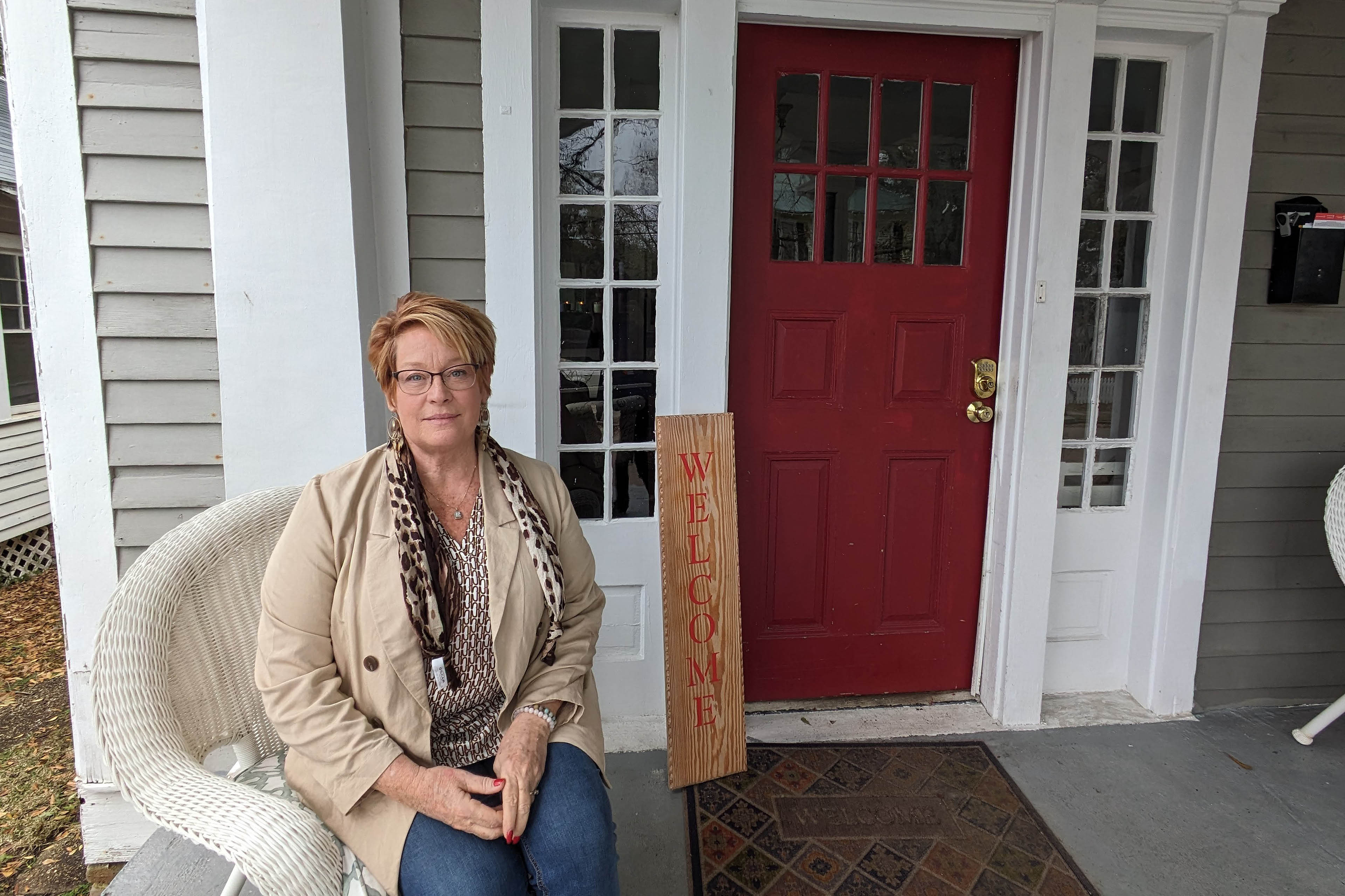 Lisa Teggart sits on the porch of a small house. Behind her is a red door and a vertical sign that reads, 