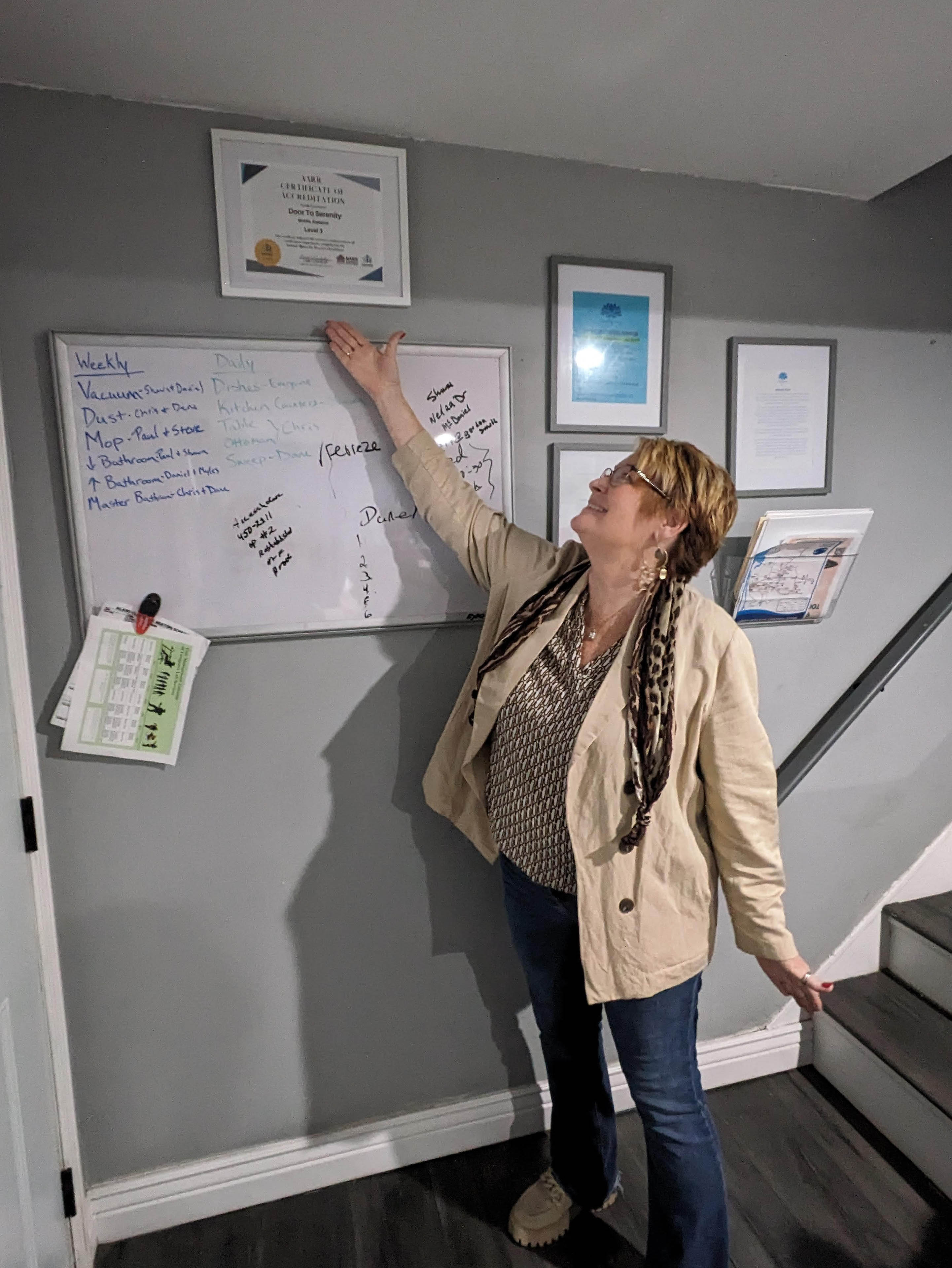 Lisa Teggart proudly points to her certification from the Alabama Alliance for Recovery Residences. The certificate hangs on a gray wall beside a staircase.