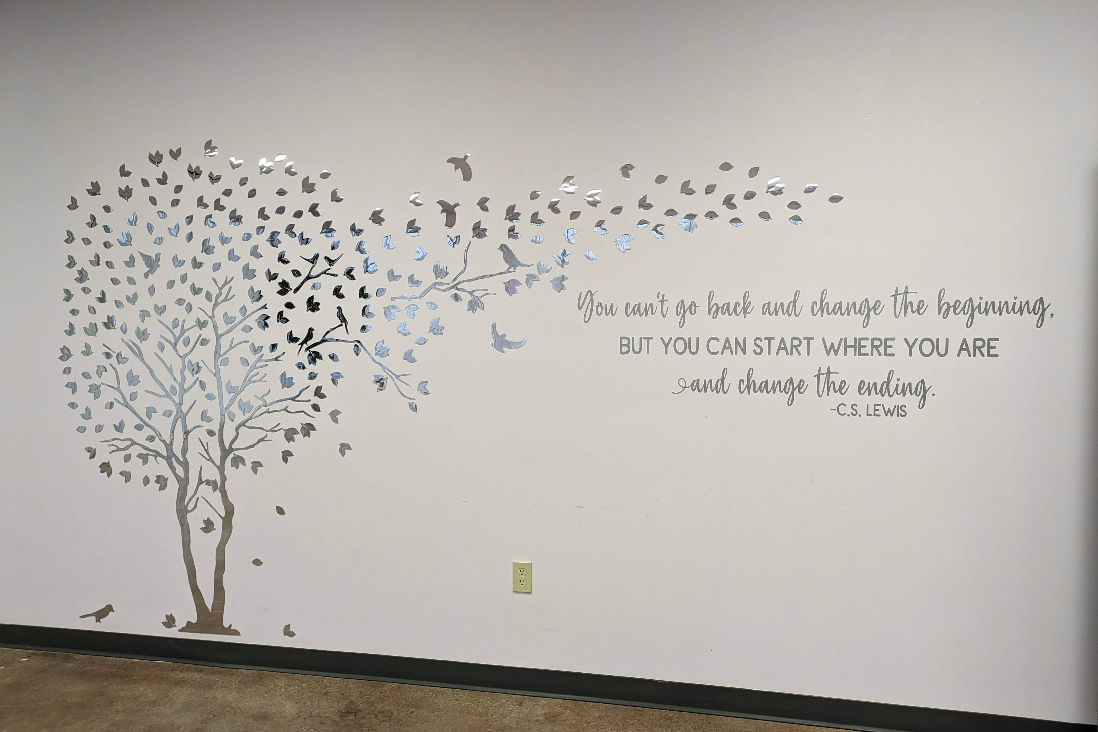 A painting of a silver tree is on a wall. Leaves blow off the tree and are accompanied by flying birds. A quote is to the left of the tree, and it reads, 