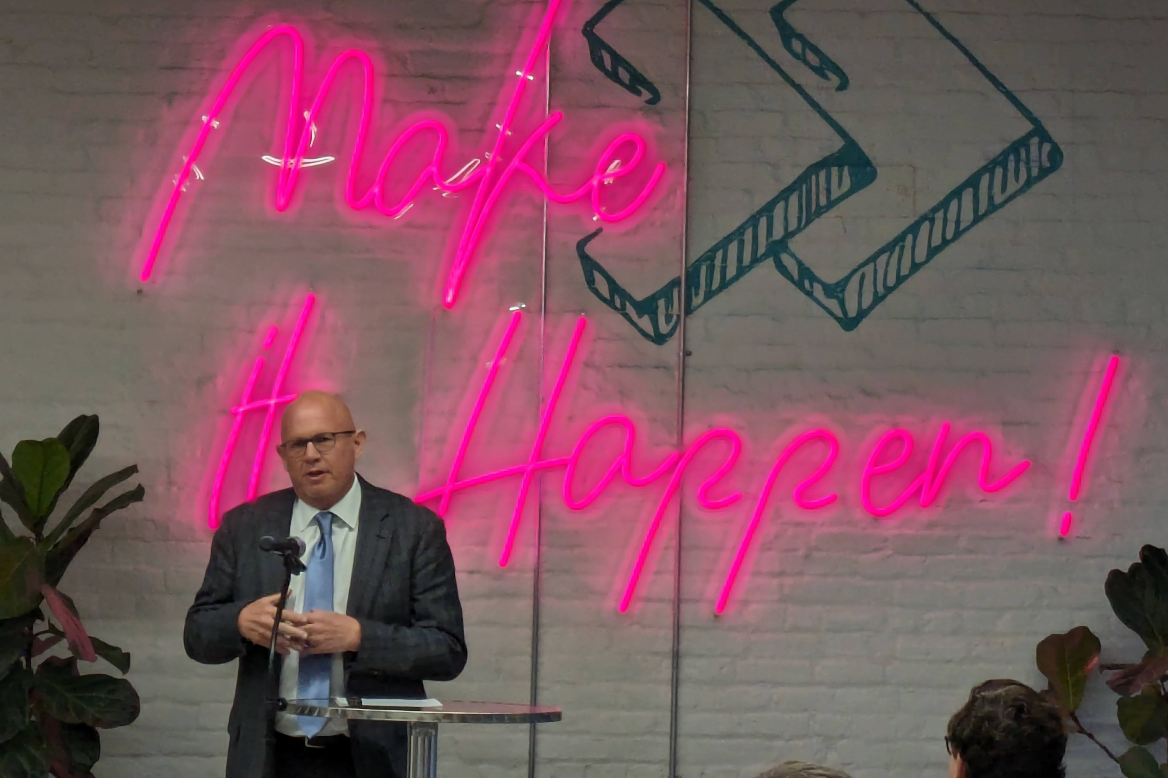 Stephen Loyd stands at a podium as he speaks at a convention. Behind him is a large, pink neon sign that reads, "Make it happen!"