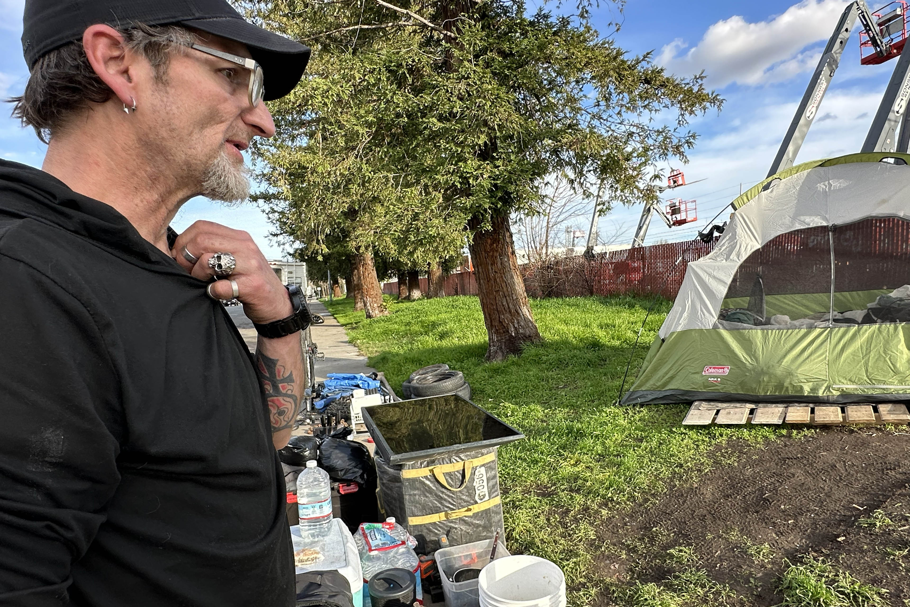 Samuel Buckles stands to the left of the image and looks at his encampment area as he prepares to leave the area. His tent and belongings are organized on the grass and sidewalk.