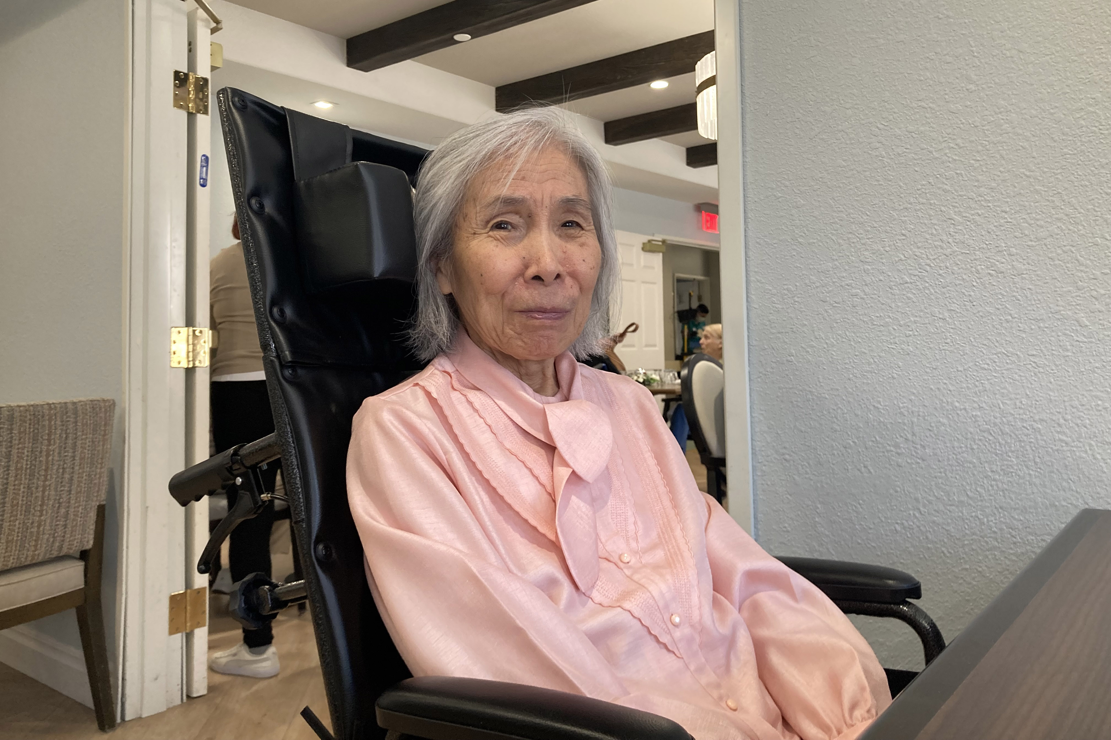 Fujiko Shinozaki, a senior woman, sits in a wheelchair and looks towards the camera. She is inside a nursing home.
