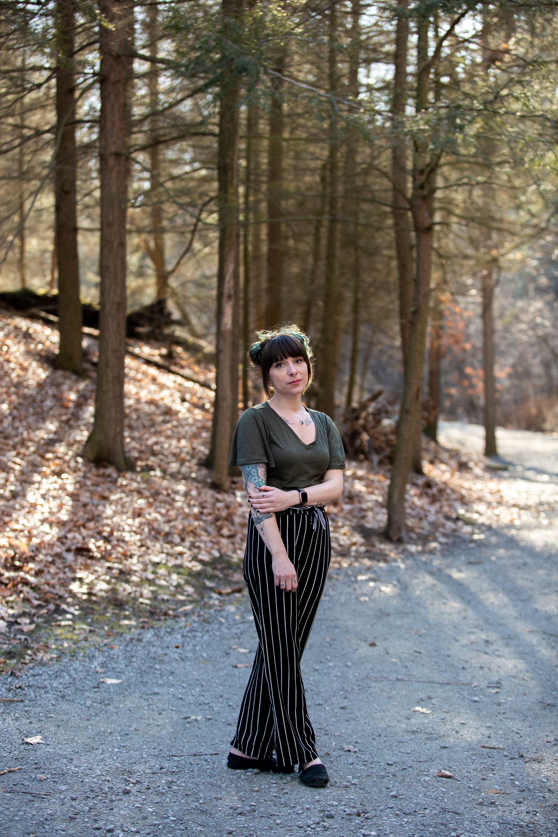 A wide shote of a woman standing outside in the woods and looking at the camera.
