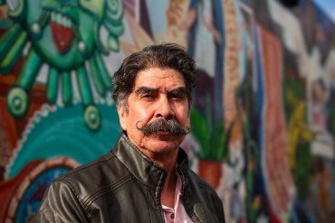 A portrait of Antonio Abundis. He stands in front of a colorful mural on a sunny day.