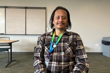 Derrick Cordero sits in a chair in the center of the photograph. He rests his hands on his knees and looks directly towards the camera with a smile.