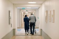 A photo of a female nurse helping an elderly man using a walker down a corridor.