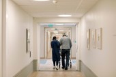 A rear view photograph of a nurse walking with senior man in a corridor at a nursing home.
