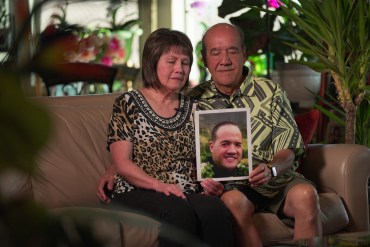 William Haleck (right) sits on a couch beside his wife, Verdell (left) with his arm around her. They both hold a photo of their son, Sheldon, in their lap and look solemnly down towards the picture.