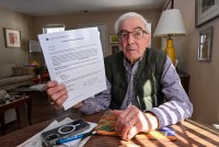 A senior man holds a letter from a Medicare provider. He is seated a table wearing glasses and a shirt and vest