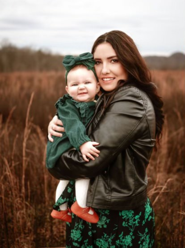Jessica Neal holds her baby in her arms in this stylized professional portrait. The baby wears a green dress with a large green bow on its head.