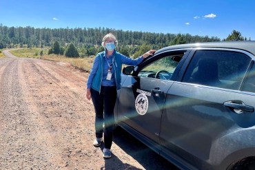 A woman in a blue shirt and mask stands in front of a car parked along a dirt road