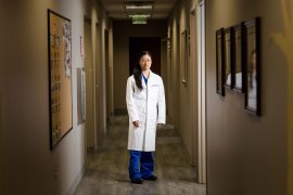 A woman with long dark hair and wearing a white doctor's coat stands in a hallway with arms by her side and looks at the camera.