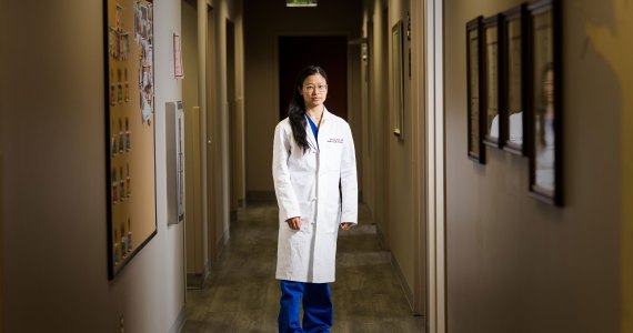 A woman with long dark hair and wearing a white doctor's coat stands in a hallway with arms by her side and looks at the camera.