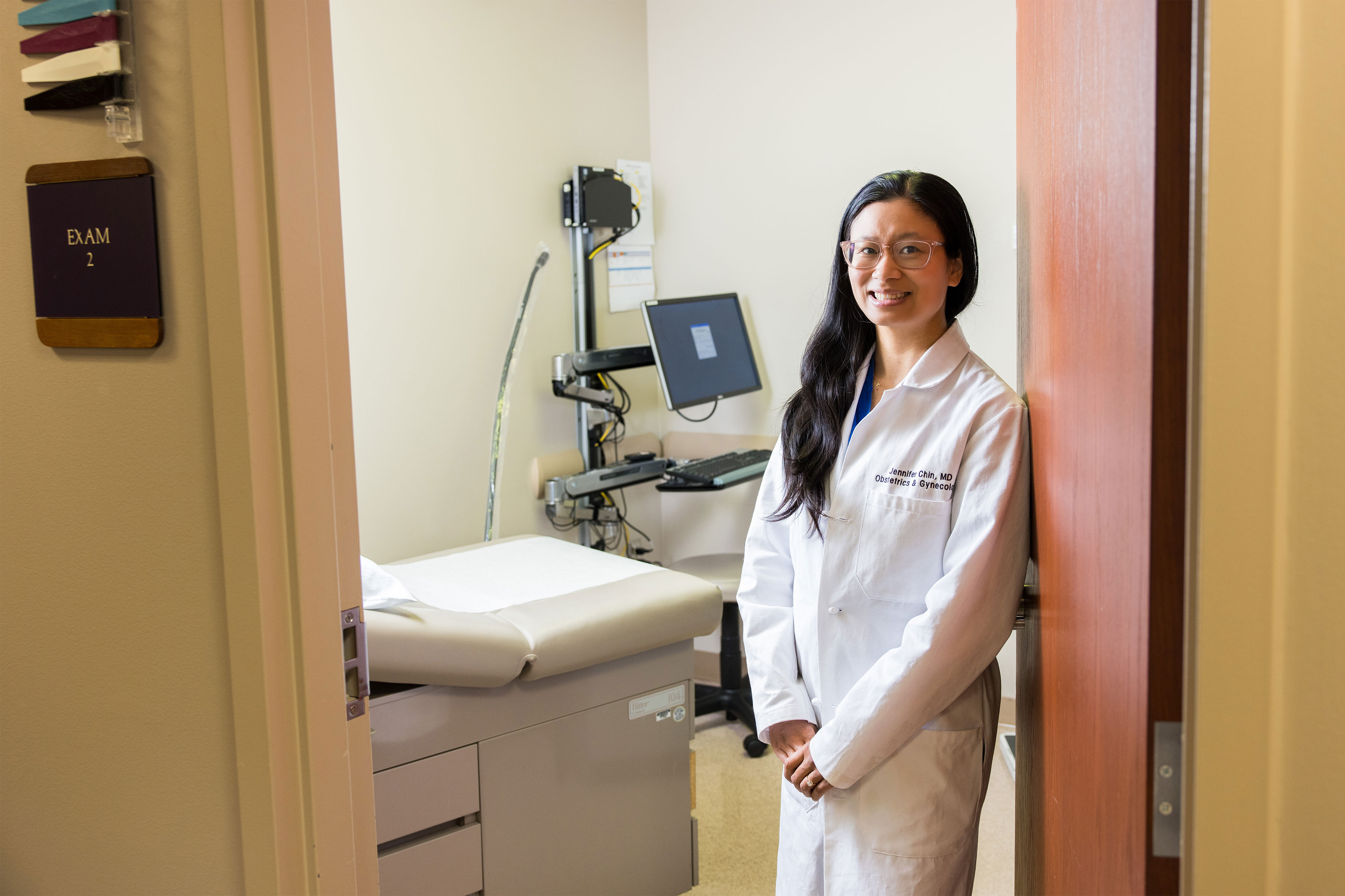 A woman with long dark hair and wearing glasses and a white doctor's coat stands just inside the doorway of an exam room. 