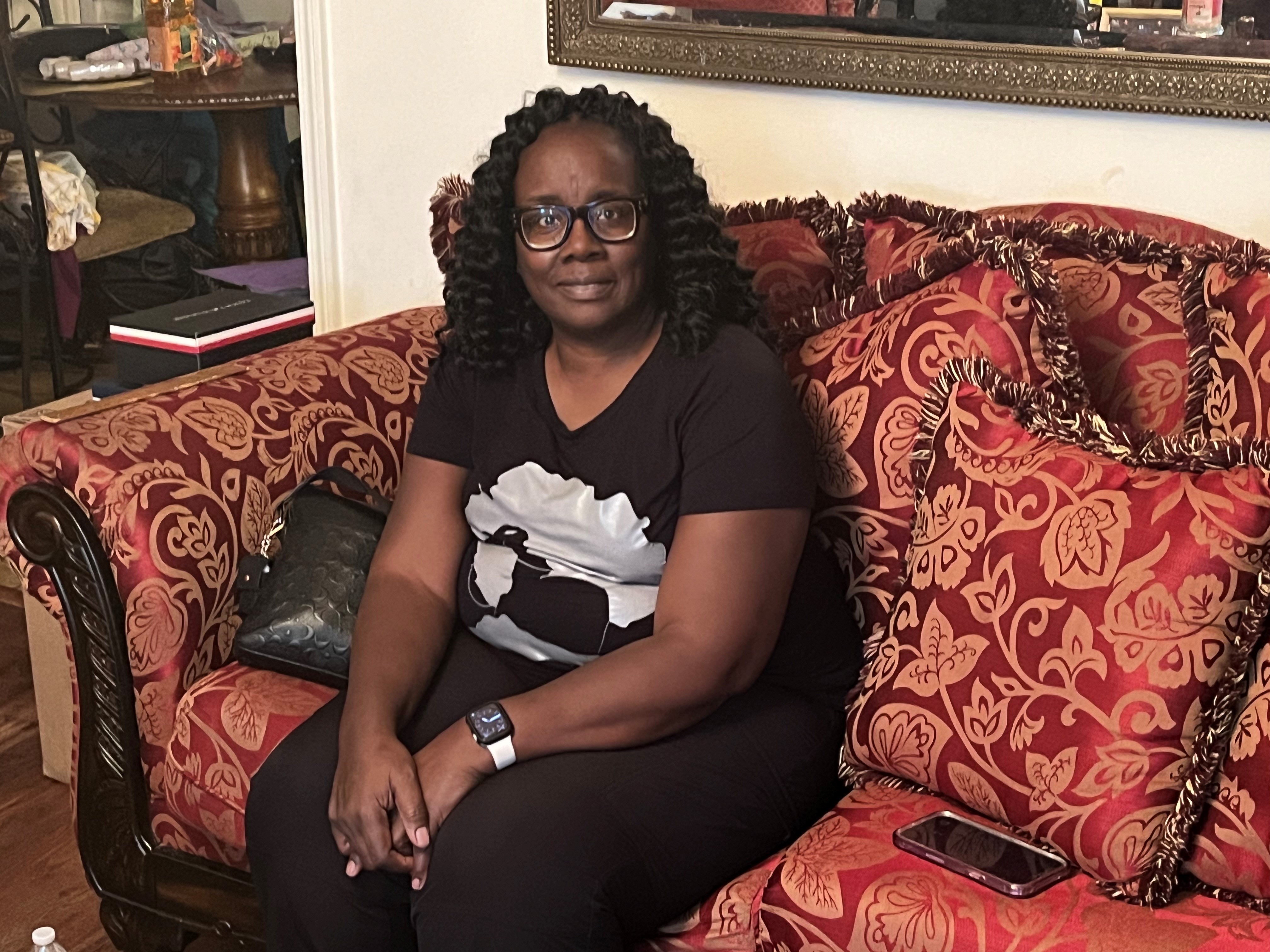 A woman in a black tshirt and glasses is seated on a red couch, looking at the camera