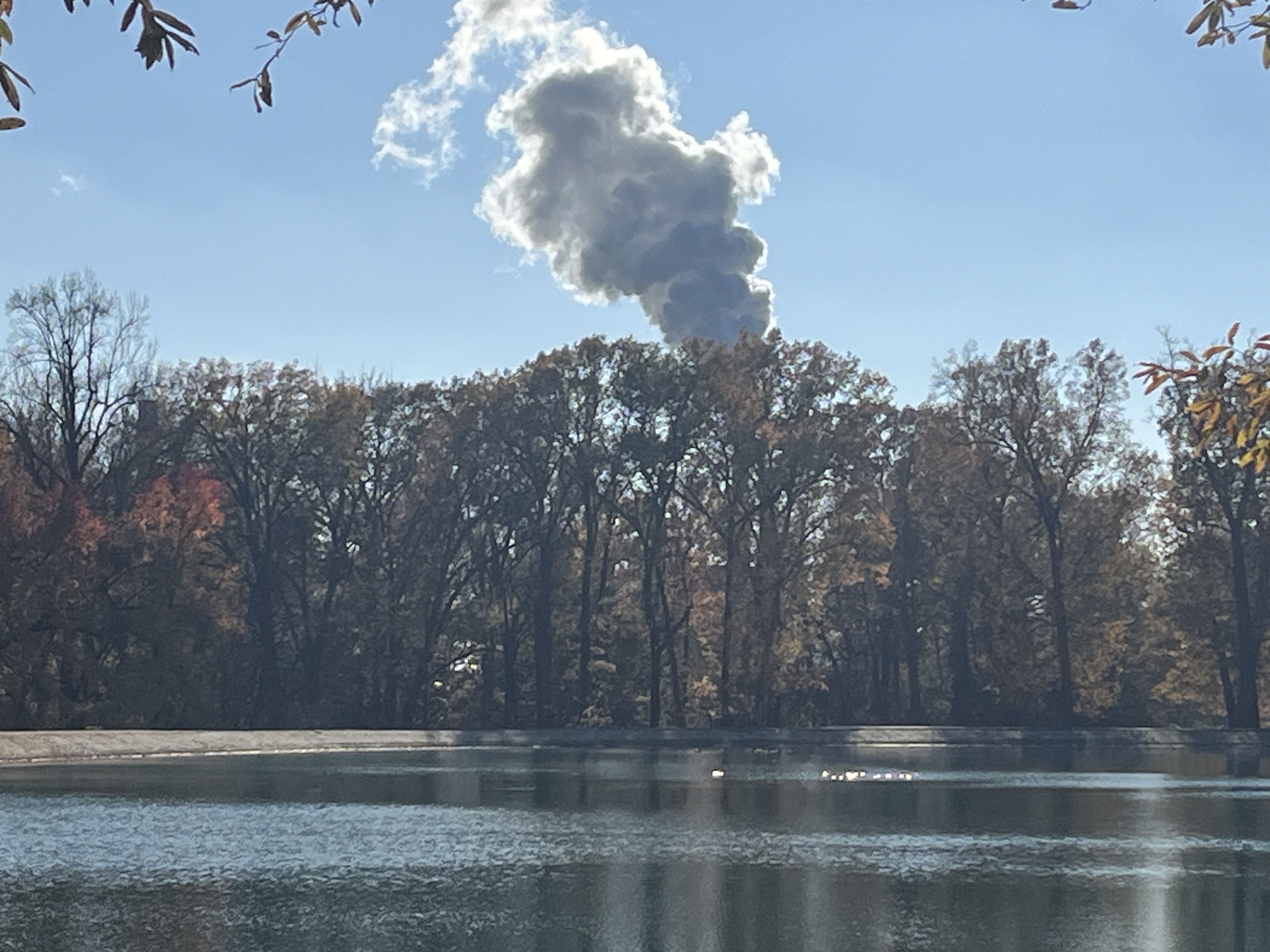 A smoke plume billows behind a row of trees that line a lake