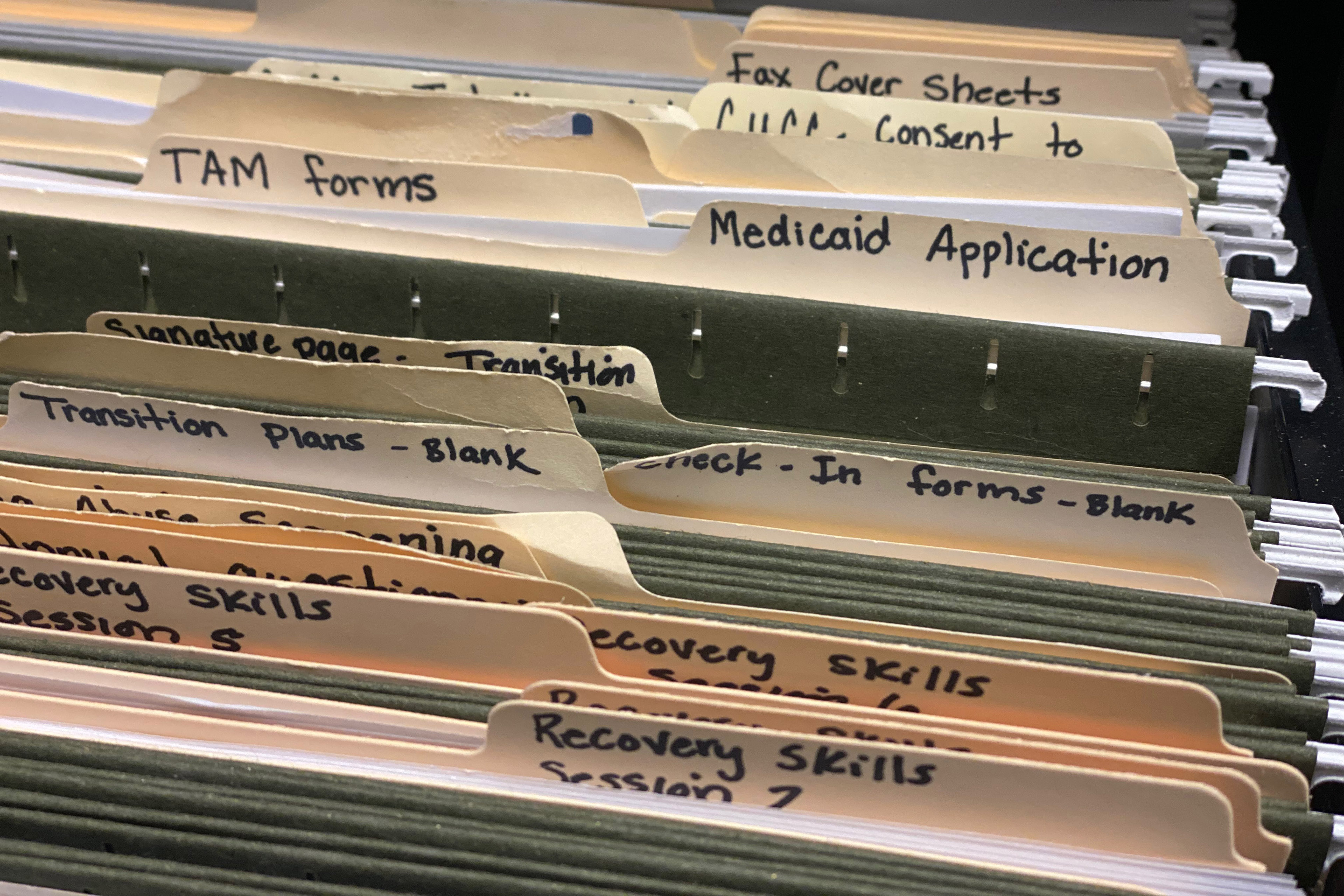 A photograph of a filing cabinet drawer. The folders are labeled in black sharpie. Some that are visible say, "TAM forms / Medicaid Application / Transition plans - Blank / Check-In forms – Blank / Recovery Skills."
