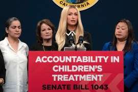 A tall, blonde woman in her early 40's stands a podium. The podium has a red sign that reads, "ACCOUNTABILITY IN CHILDREN'S TREATMENT / SENATE BILL 1043"