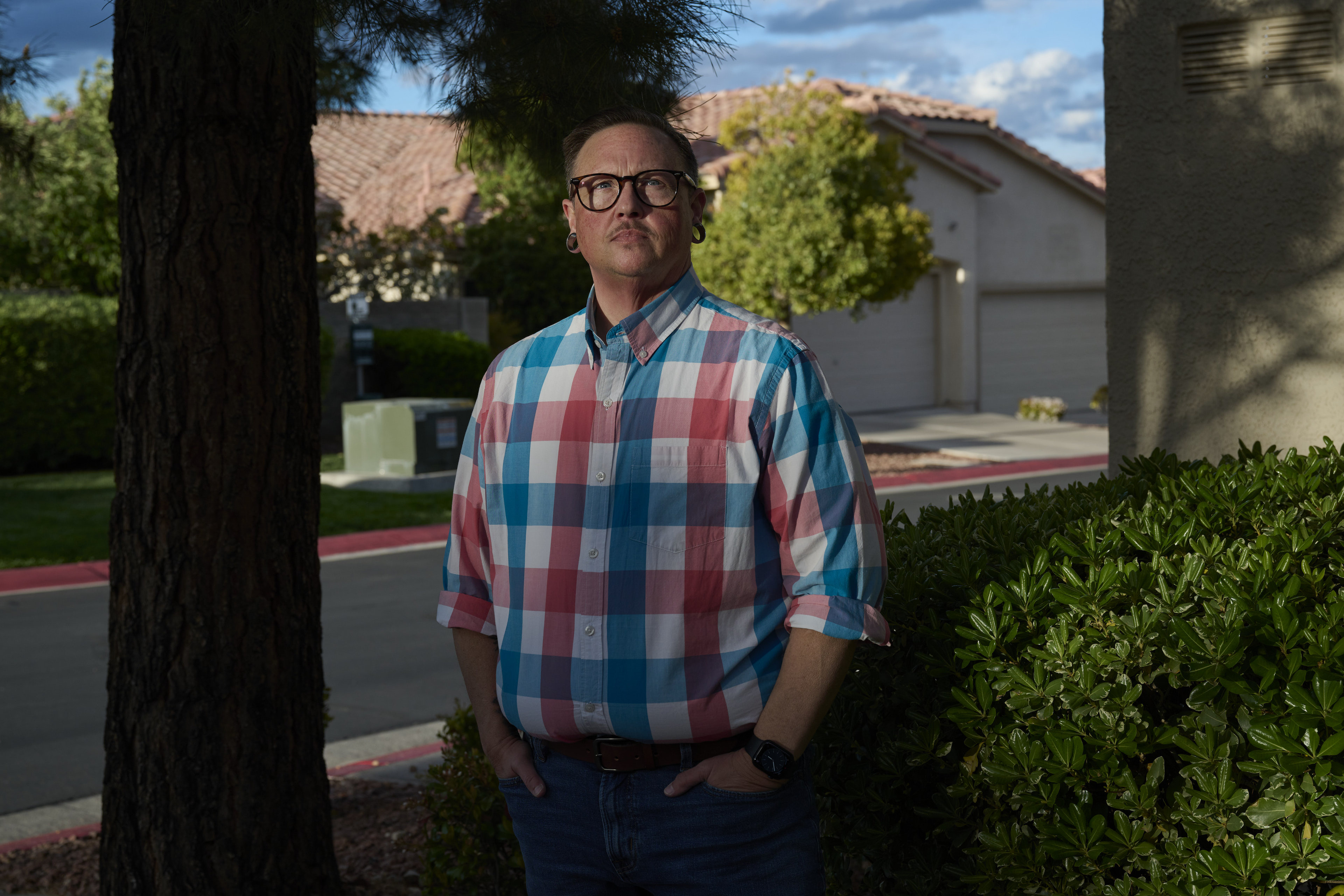 A man in a plaid shirt and glasses stands in front of a street of houses