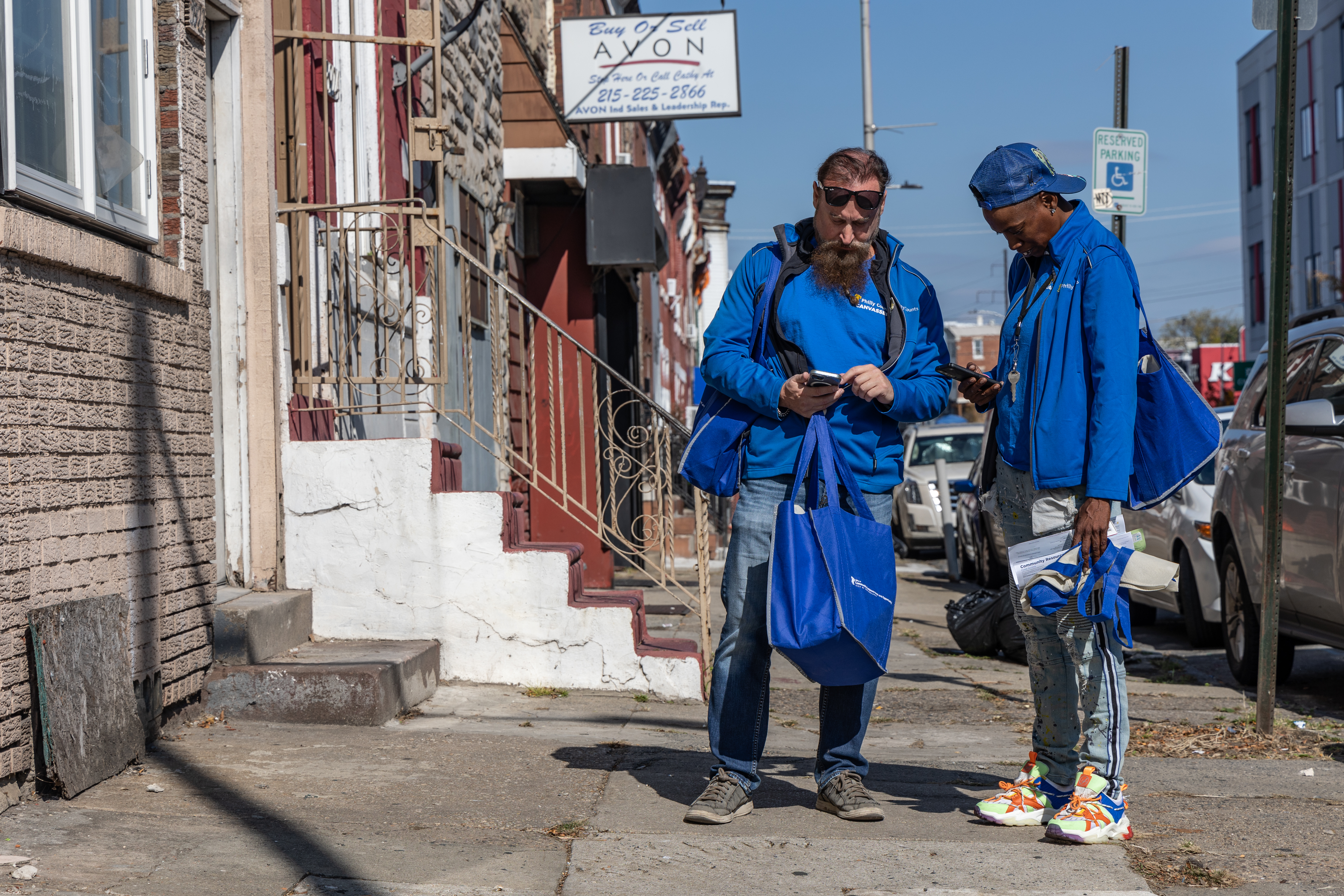 Two people in matching blue sweat jackets stand on a sidewalk looking at their phones.