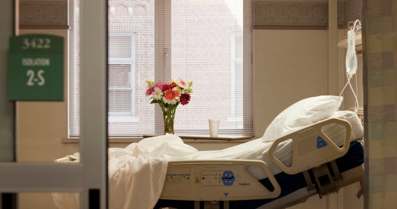 A photo showing a vacant hospital room. The bed is empty and there are flowers on the window sill.