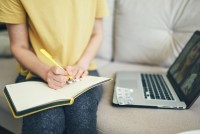 A close up photo of a person taking notes in a journal while on a telehealth call. Their laptop is on the couch beside them.