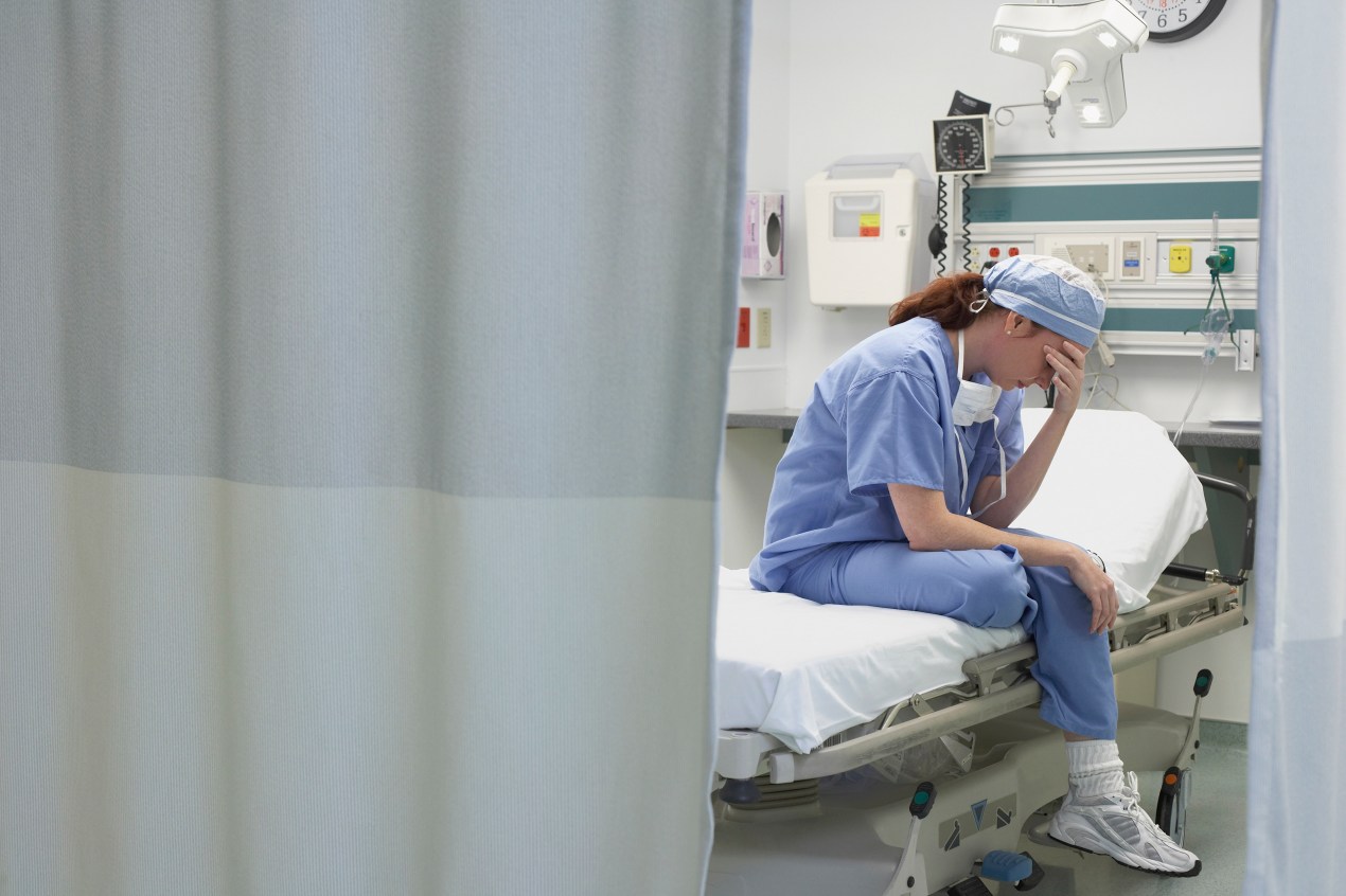 A female doctor sits with her head in hand hand on an empty hospital bed.