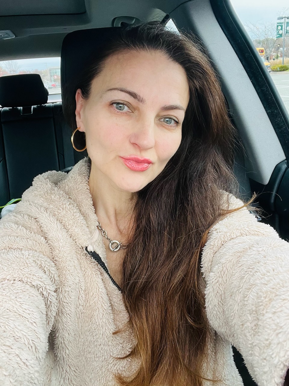 A woman with long brown hair takes a selfie while sitting in a car.