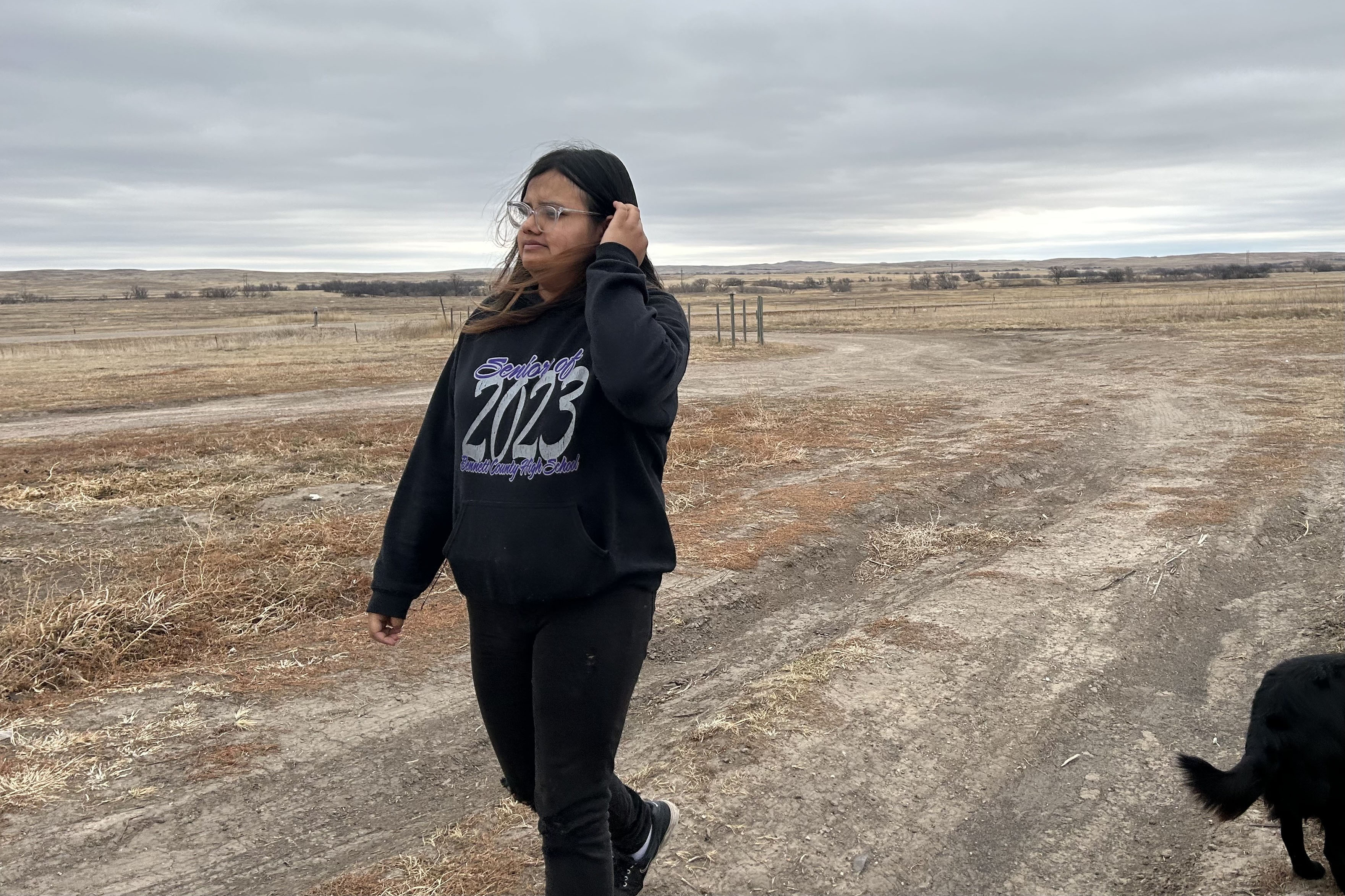 Katherine Goodlow crosses her family’s driveway in Hisle on the Pine Ridge Reservation in South Dakota. The sky is overcast and the landscape shows a dirt road and dry, grassy fields.