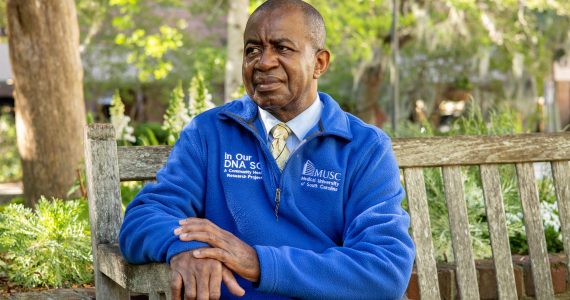 A photo of Lee Moultrie sitting on a bench outside in a blue pullover.