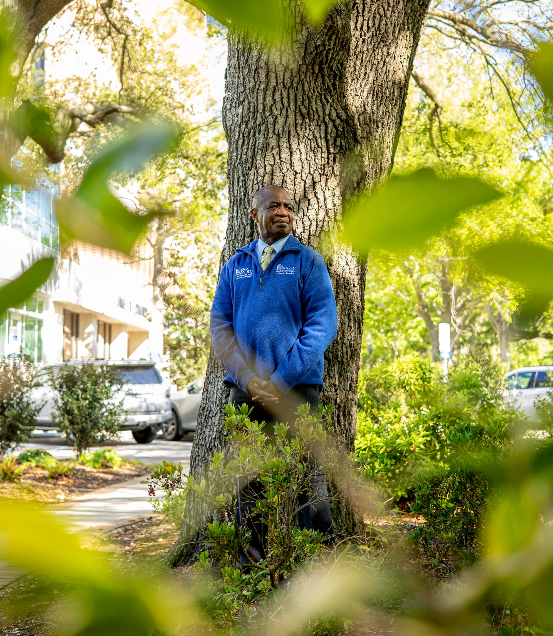 A portrait of a black man standing outside. Blurred foliage in the foreground frames him.