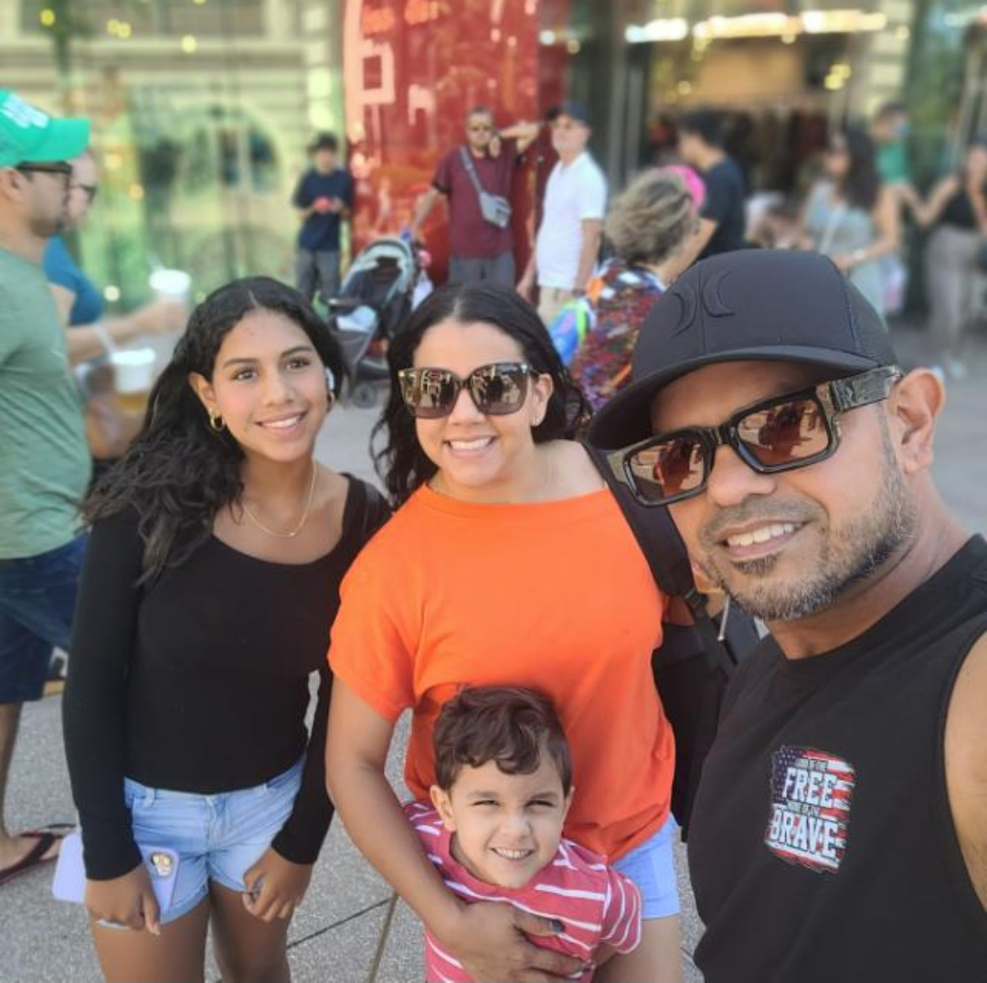 A man takes a selfie of himself, a woman, and two children on a busy sidewalk.