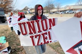 A photo of a man in a line with other protesters. He is holding a sign that reads, "Unionize!"