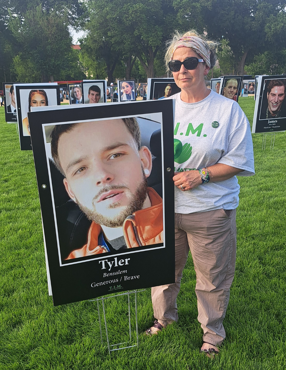 Susan Ousterman stands in a grassy area holding a poster-size photo of her son, Tyler Cordeiro. Behind her are more rows of poster-sized photographs of others who have passed away.