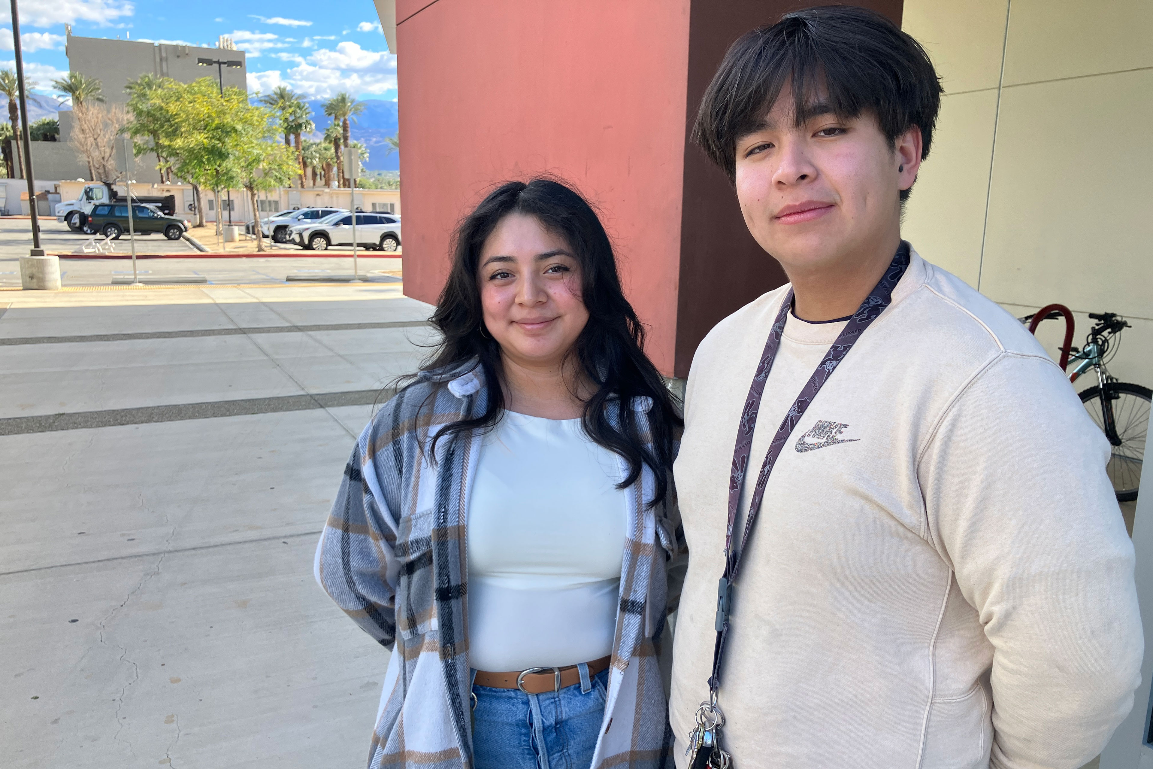 Two young adults stand on a sidewalk just outside a building. Both have dark hair and are smiling at the camera.