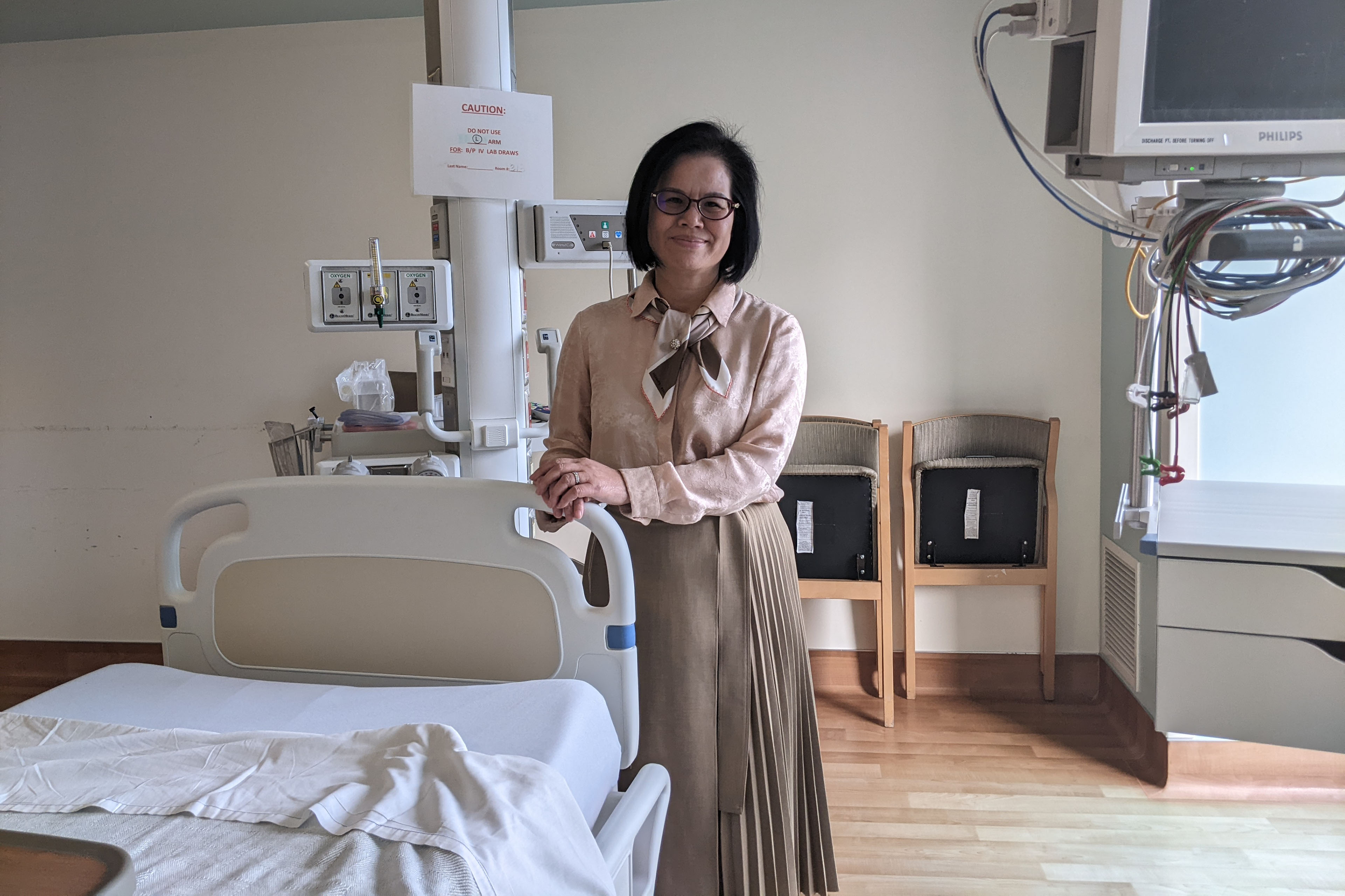 Jian Zhang stands beside a hospital bed and smiles looking toward the camera.