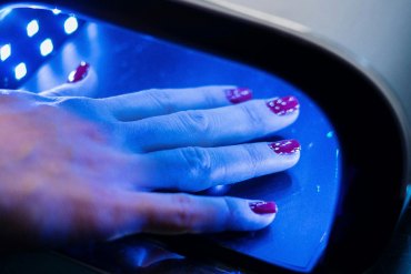 A person with painted nails holds their hand under machine emitting a blue light.