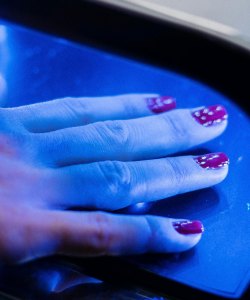 A person with painted nails holds their hand under machine emitting a blue light.