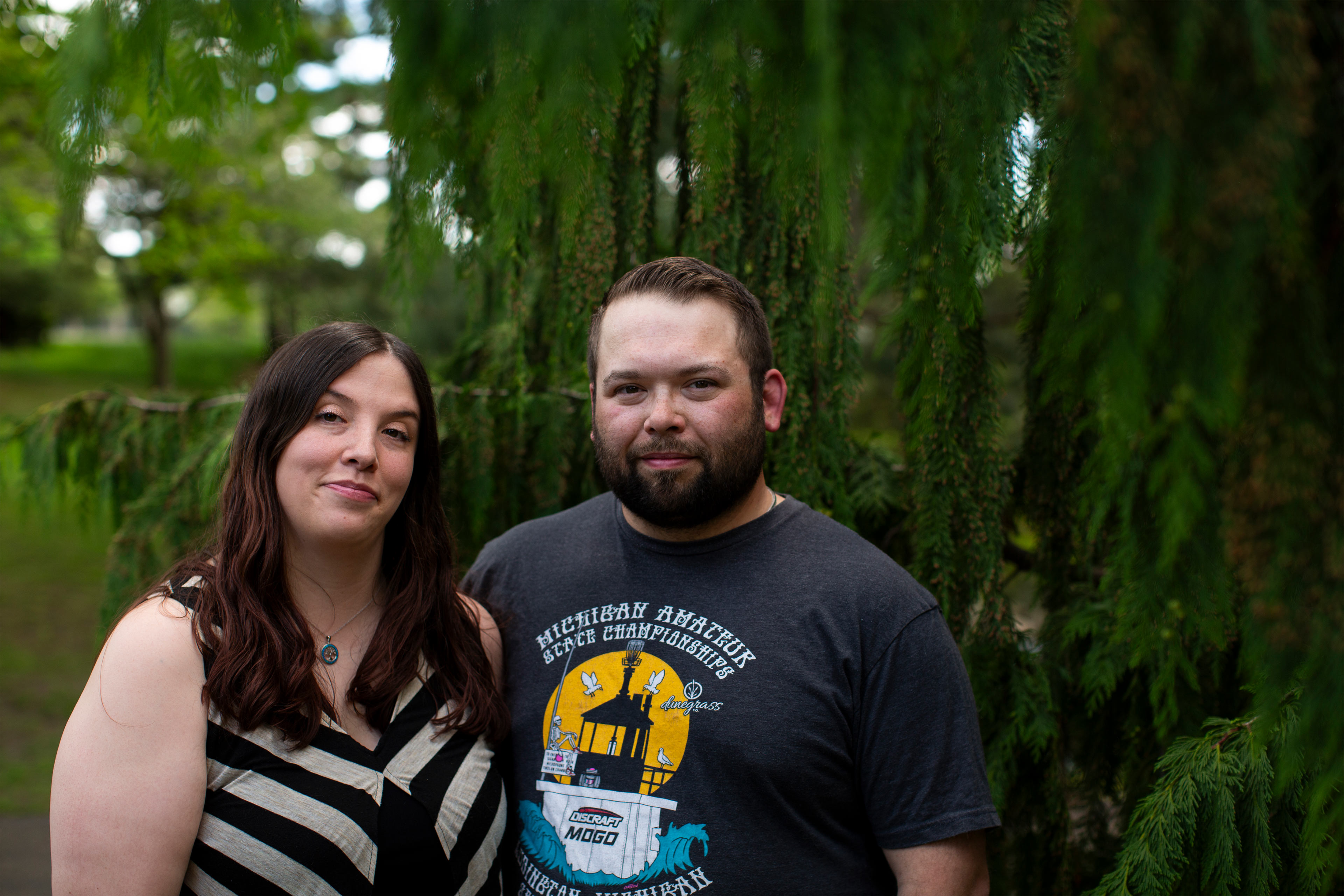 A man and a woman stand with an arm around the other outside by a tree and look at the camera.