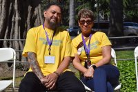 A photo of a man and a woman sitting next to each other outside. They both are wearing yellow shirts that read, "People with disabilities deserve services."