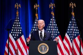 A photo of President Biden speaking at a podium with four American flags behind him.